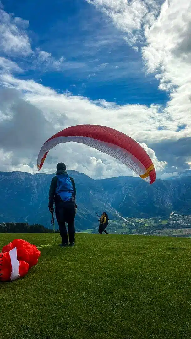 Gleitschirmpilot beim Aufziehen des rot-weißen Schirms am Startplatz, im Hintergrund Berge und Tal