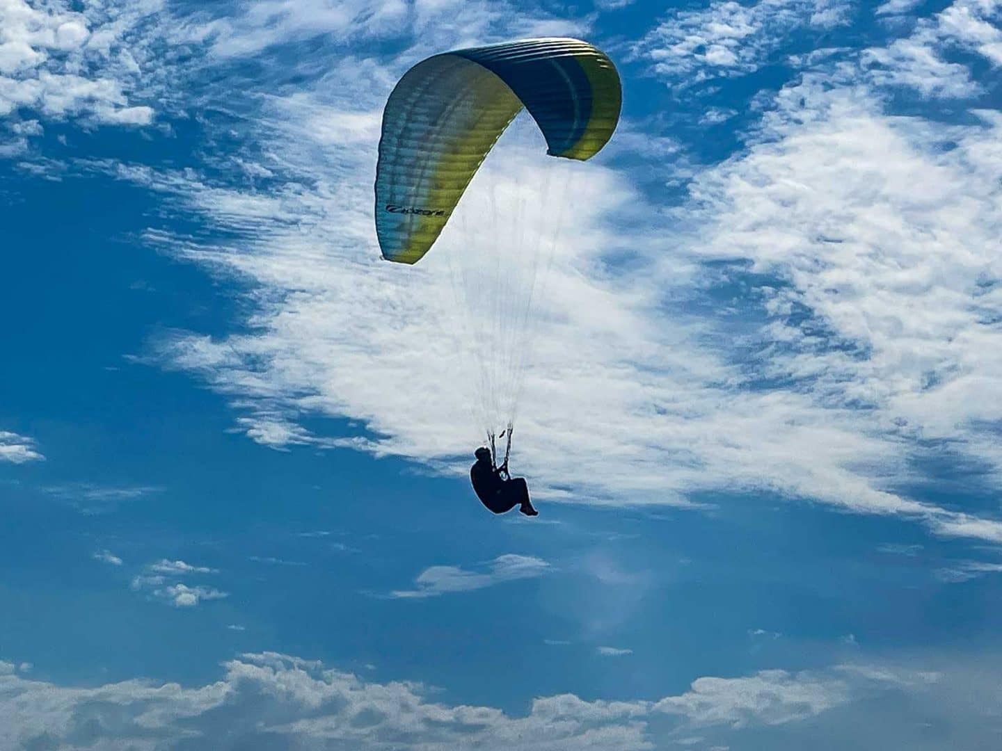 Gleitschirmpilot im Gurtzeug unter gelbgrüner Kappe vor Wolkenhimmel