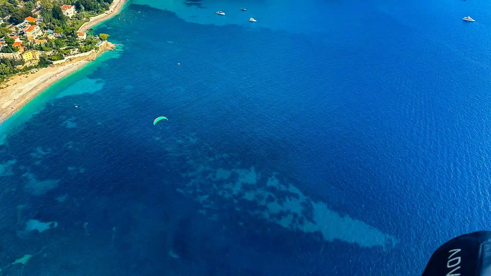Blick von oben auf das tiefblaue Mittelmeer, am Rand ein Sandstrand und ein Gleitschirm