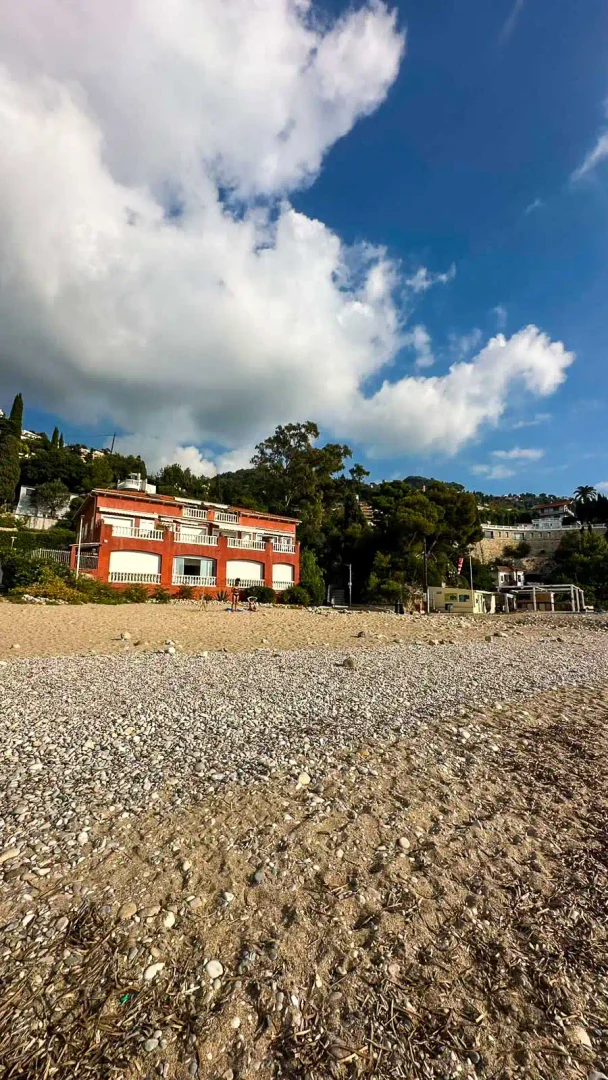 Kieselstrand mit rotem Gebäude und Bäumen unter blauem Himmel